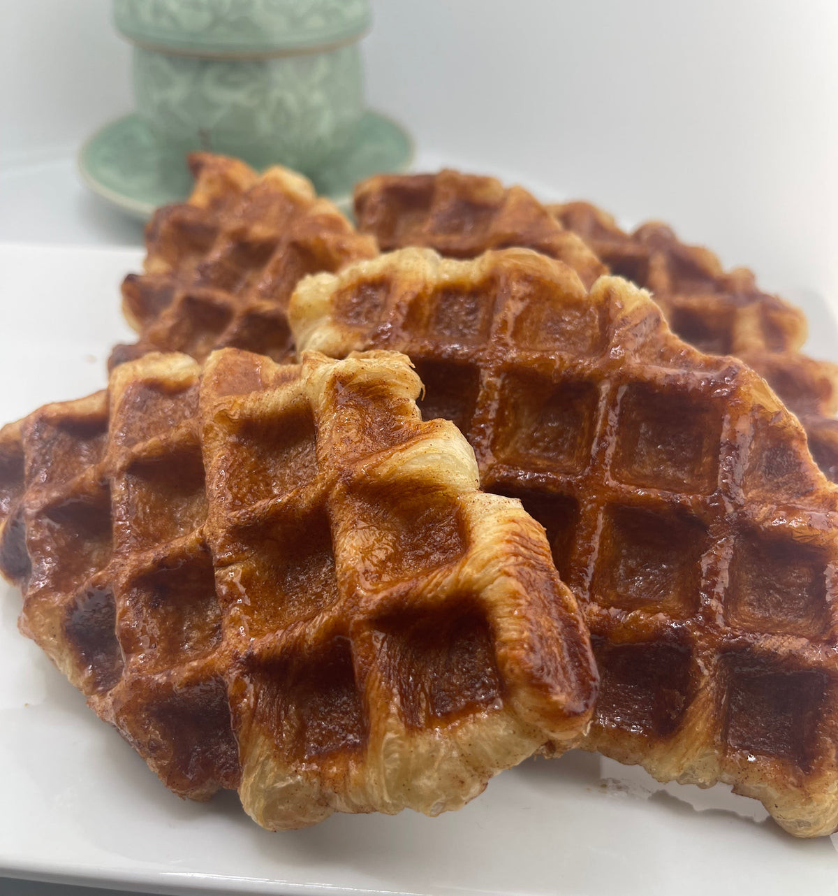 Golden-brown baked waffles stacked on a white plate, with a small pale green decorative container blurred in the background.