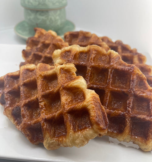 Golden-brown baked waffles stacked on a white plate, with a small pale green decorative container blurred in the background.